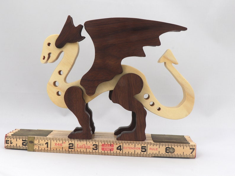 Handmade wooden dragon figurine made from contrasting hardwoods with a light natural wood body and head paired with rich dark reddish-brown wings, legs, and horns. Displayed on a wooden carpenters rule for scale.