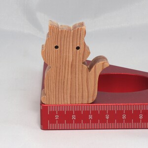 May include: A wooden cat figurine with a natural wood finish, sitting on a red protractor with a ruler marked in inches.