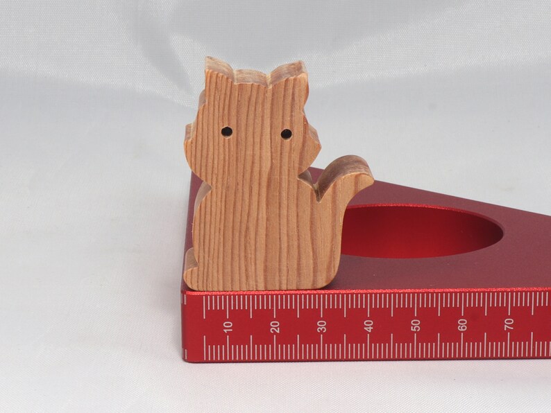 May include: A wooden cat figurine with a natural wood finish, sitting on a red protractor with a ruler marked in inches.