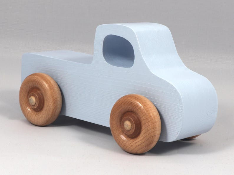 May include: A light blue wooden toy truck with natural wood wheels. The truck has a classic pickup design with a cab and a cargo bed. The wheels are round and made of light-colored wood. The toy is painted in a soft, pastel blue.