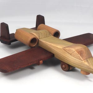 May include: A wooden toy airplane with a natural wood finish and dark brown wings and tail. The toy features two cylindrical engine housings and small wheels. The plane is designed with a detailed cockpit.
