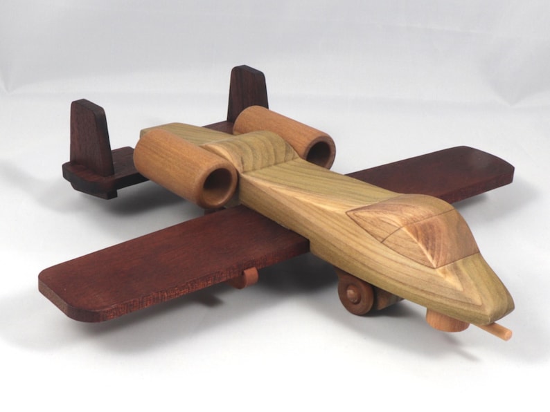 May include: A wooden toy airplane with a natural wood finish and dark brown wings and tail. The toy features two cylindrical engine housings and small wheels. The plane is designed with a detailed cockpit.