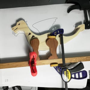 Wooden dragon under assembly in the workshop. Red and blue clamps hold two dark wooden legs against its light-colored body, which rests over a paper pattern showing the wing and remaining leg outlines.