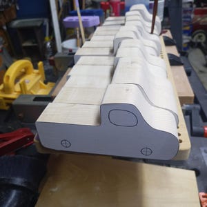 A row of unfinished wooden toy pickup bodies on a workshop bench, with paper patterns showing the cab windows and wheel positions.