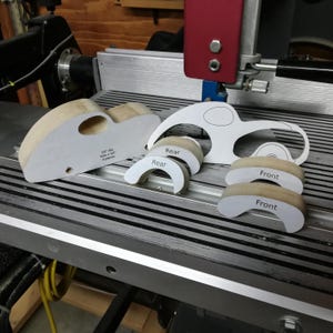 Unassembled wooden toy car parts arranged on a Shopsmith bandsaw table. The cut pine body on the left has an oval window opening and drilled axle holes, with its paper pattern still attached. Four curved fat-fender pieces sit in front, two labeled “Rear” and two labeled “Front.” A paper car template with the fender areas cut away stands behind the pieces; it is used to precisely locate the fenders for assembly.