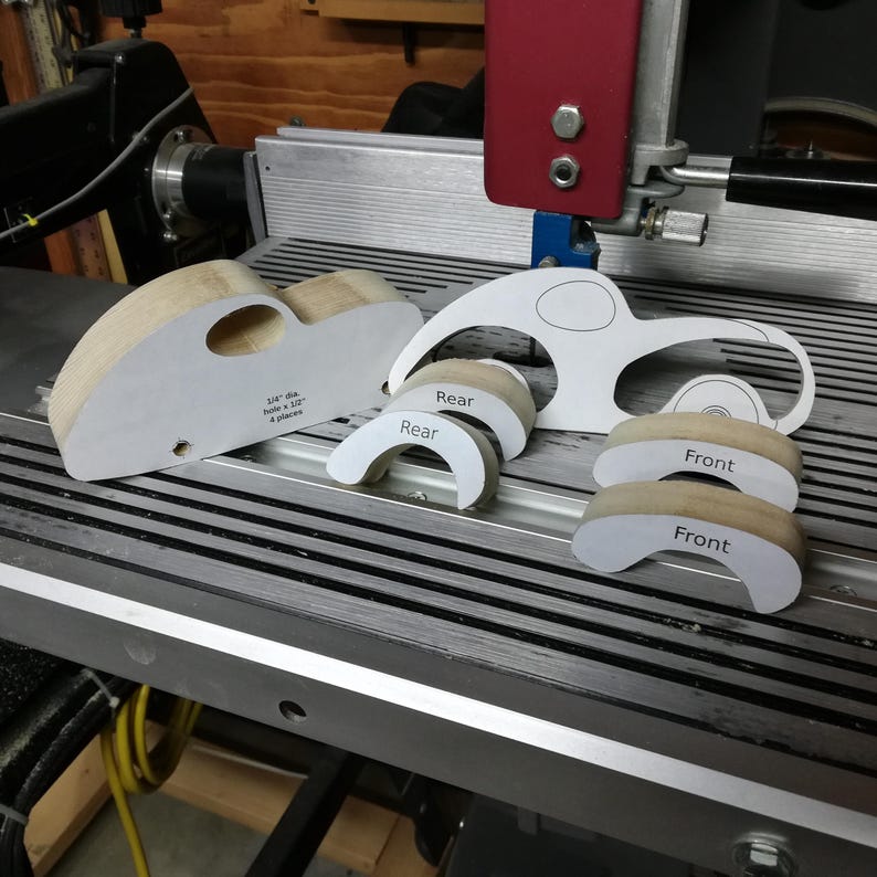 Unassembled wooden toy car parts arranged on a Shopsmith bandsaw table. The cut pine body on the left has an oval window opening and drilled axle holes, with its paper pattern still attached. Four curved fat-fender pieces sit in front, two labeled “Rear” and two labeled “Front.” A paper car template with the fender areas cut away stands behind the pieces; it is used to precisely locate the fenders for assembly.