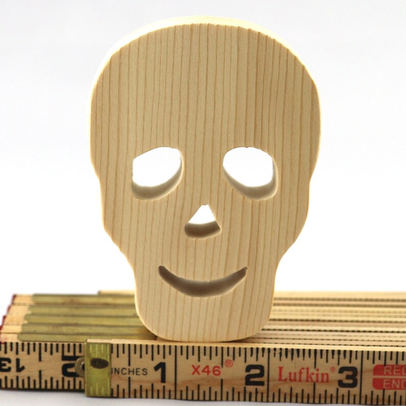 A handmade, unfinished light-wood Halloween skull cutout features carved eye sockets, a nose, and a smiling mouth, standing atop a folding wooden carpenter's rule for scale.