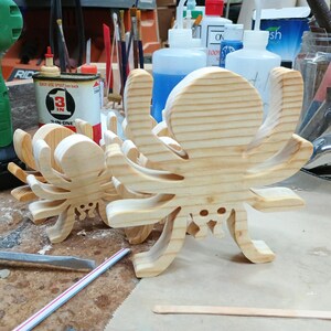 May include: Two light-colored wooden spider cutouts featuring a natural grain pattern. Each spider has a rounded body, eight curved legs, and two small circular cutouts near the bottom representing eyes, standing upright on a flat surface.