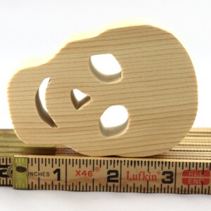 A handmade, unfinished light wood Halloween skull cutout features carved eye sockets, a nose, and a smiling mouth on a white background, pictured lying on its side atop a folding wooden carpenter's rule for scale.