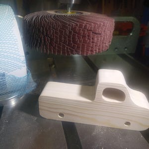 Unfinished wooden toy pickup body beside a flap sanding wheel, showing its smoothed edges, cutout cab window, and drilled axle holes.
