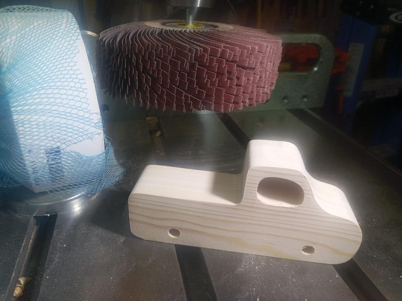 Unfinished wooden toy pickup body beside a flap sanding wheel, showing its smoothed edges, cutout cab window, and drilled axle holes.