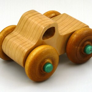 A handmade wooden toy monster truck with a natural wood body and four large wheels. The body and wheels are finished with amber shellac, and the hubs are painted with metallac emerald green acrylic paint. The toy is designed with a simple, classic aesthetic.