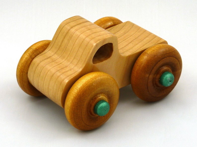A handmade wooden toy monster truck with a natural wood body and four large wheels. The body and wheels are finished with amber shellac, and the hubs are painted with metallac emerald green acrylic paint. The toy is designed with a simple, classic aesthetic.