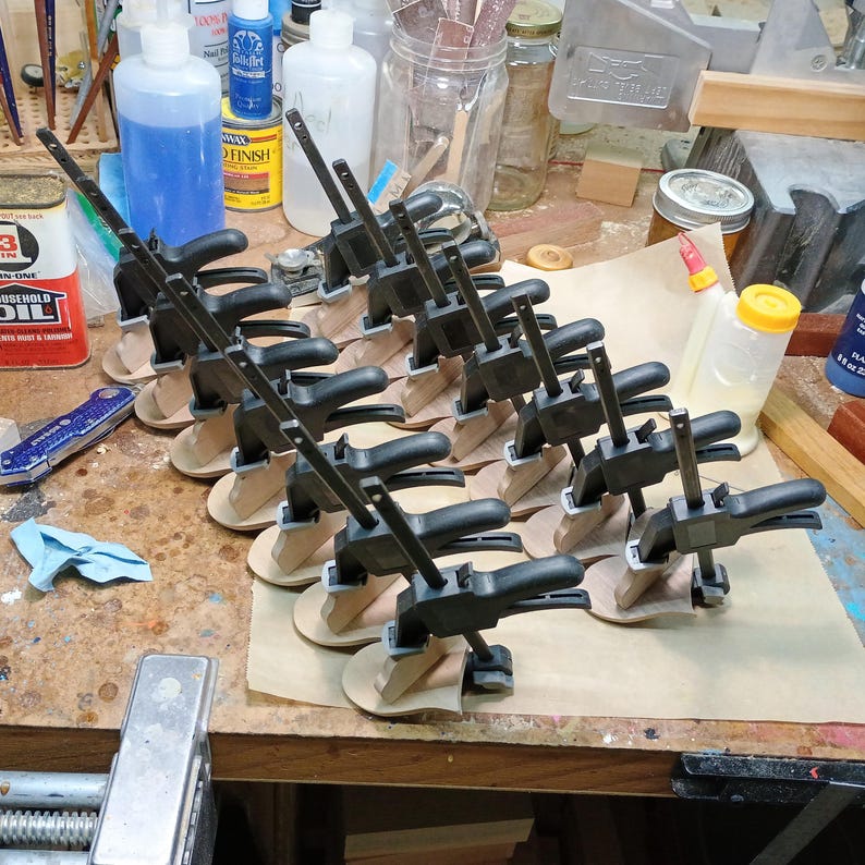 May include: A collection of black plastic bar clamps secured onto small, light-colored wooden pieces arranged in rows on a sheet of parchment paper. Each clamp features a sliding jaw mechanism and a handle, positioned to hold the wood components firmly.