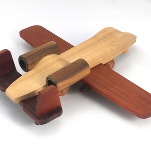 May include: A wooden toy airplane with a natural wood body, dark brown accents, and reddish-brown wings and tail. The toy has a classic design with a propeller and small wheels, perfect for imaginative play.