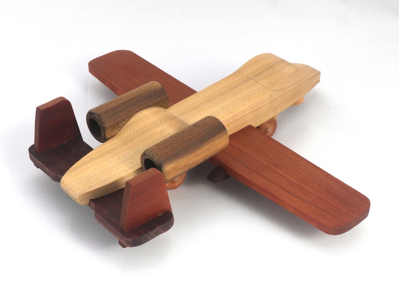 May include: A wooden toy airplane with a natural wood body, dark brown accents, and reddish-brown wings and tail. The toy has a classic design with a propeller and small wheels, perfect for imaginative play.