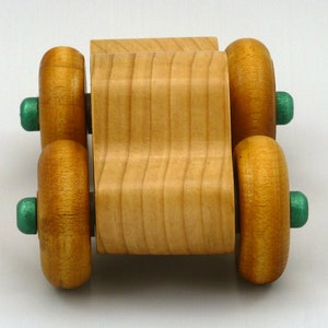 A handmade wooden toy monster truck with a natural wood body and four large wheels. The body and wheels are finished with amber shellac, and the hubs are painted with metallac emerald green acrylic paint. The toy is designed with a simple, classic aesthetic.