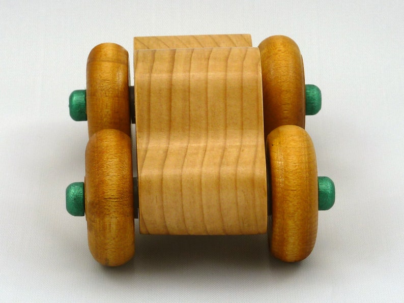 A handmade wooden toy monster truck with a natural wood body and four large wheels. The body and wheels are finished with amber shellac, and the hubs are painted with metallac emerald green acrylic paint. The toy is designed with a simple, classic aesthetic.