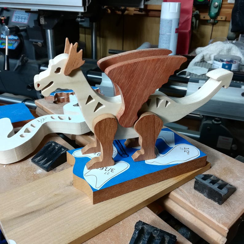 Wooden dragon figurine standing on a workshop bench, with a pale body and curled tail, brown wings, horns, and legs. Cut wooden pieces and paper patterns lie beside it.