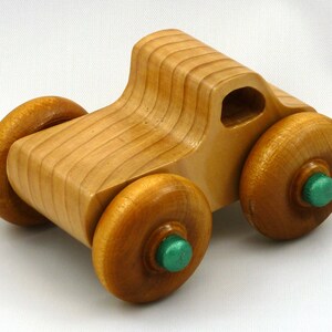 A handmade wooden toy monster truck with a natural wood body and four large wheels. The body and wheels are finished with amber shellac, and the hubs are painted with metallac emerald green acrylic paint. The toy is designed with a simple, classic aesthetic.