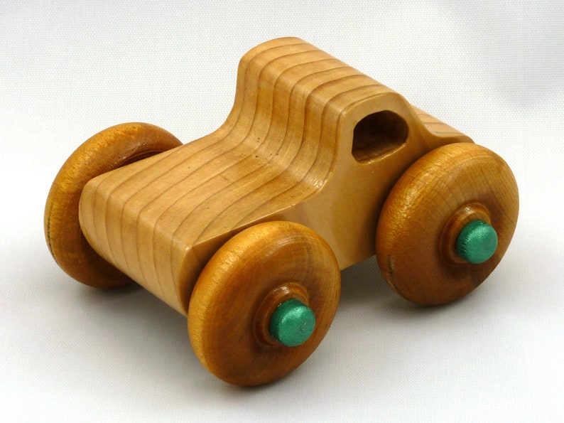 A handmade wooden toy monster truck with a natural wood body and four large wheels. The body and wheels are finished with amber shellac, and the hubs are painted with metallac emerald green acrylic paint. The toy is designed with a simple, classic aesthetic.