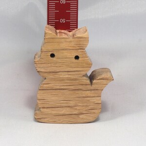 May include: A wooden cat figurine, carved from light brown wood with a natural grain pattern. The cat is sitting with its tail curled around its body. The figurine has two small holes for eyes.