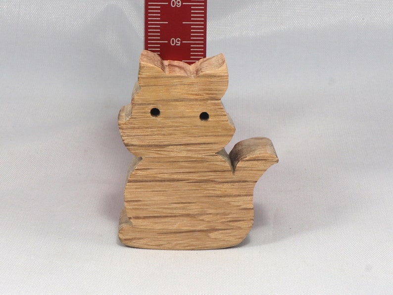 May include: A wooden cat figurine, carved from light brown wood with a natural grain pattern. The cat is sitting with its tail curled around its body. The figurine has two small holes for eyes.