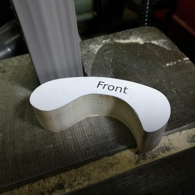 Close-up of an unfinished wooden front fender resting on a belt sander table beside the vertical sanding belt. A white paper pattern labeled “Front” remains attached to its face. The thick, curved piece has rounded ends and an arched wheel opening, forming part of the wooden toy car’s fat-fender design. Sawdust collects along the table’s grooves and edges.