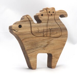 A freestanding wooden puzzle cut in the shape of a large cat standing on four legs. Two smaller kitten figures nest on the large cat’s back and interlock as puzzle pieces. Simple drilled holes form the eyes and nose on each cat, with a shallow cut line for the mouth on the large cat. The completed puzzle stands upright on a plain white surface. Handmade by Odin’s Toy Factory In Tallahassee, Florida, USA.