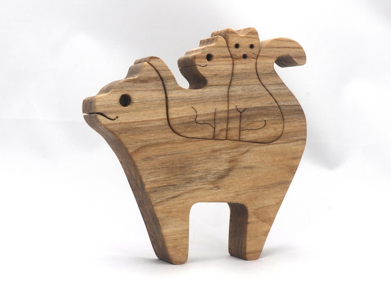 A freestanding wooden puzzle cut in the shape of a large cat standing on four legs. Two smaller kitten figures nest on the large cat’s back and interlock as puzzle pieces. Simple drilled holes form the eyes and nose on each cat, with a shallow cut line for the mouth on the large cat. The completed puzzle stands upright on a plain white surface. Handmade by Odin’s Toy Factory In Tallahassee, Florida, USA.