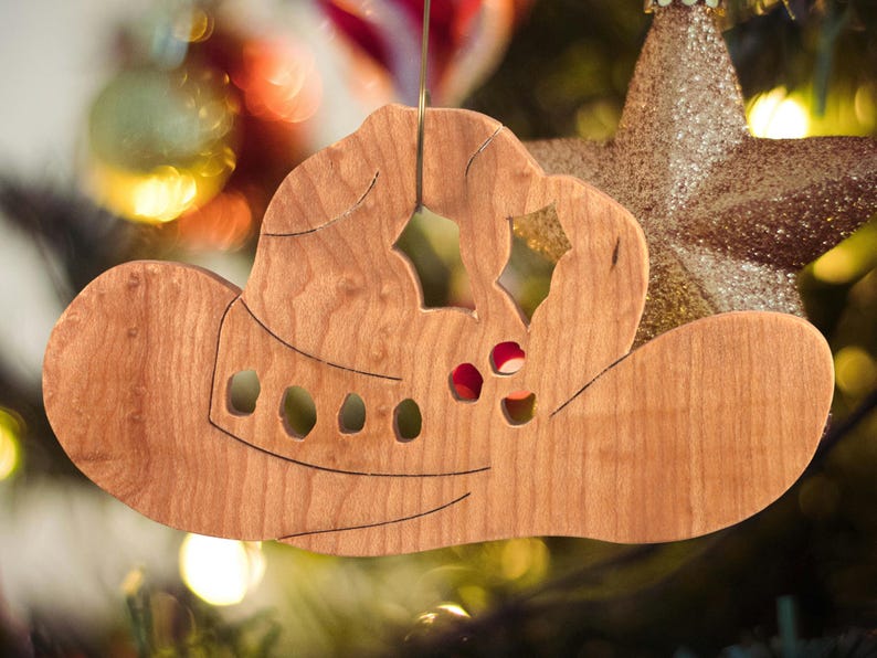 Handmade wooden Christmas tree ornament shaped like a cowboy hat, featuring a sprig of holly tucked into the hat band, star-shaped leaf cutouts, and round berry accents.