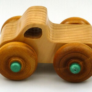 A handmade wooden toy monster truck with a natural wood body and four large wheels. The body and wheels are finished with amber shellac, and the hubs are painted with metallac emerald green acrylic paint. The toy is designed with a simple, classic aesthetic.