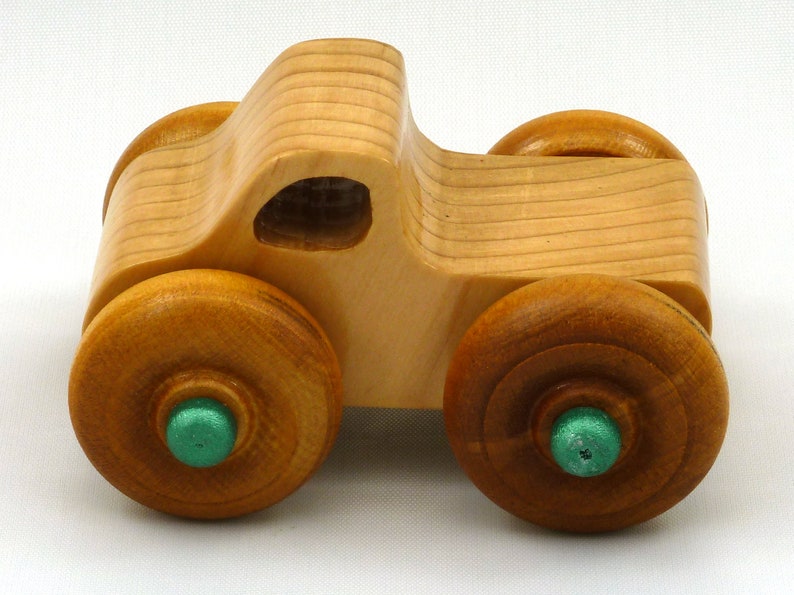 A handmade wooden toy monster truck with a natural wood body and four large wheels. The body and wheels are finished with amber shellac, and the hubs are painted with metallac emerald green acrylic paint. The toy is designed with a simple, classic aesthetic.