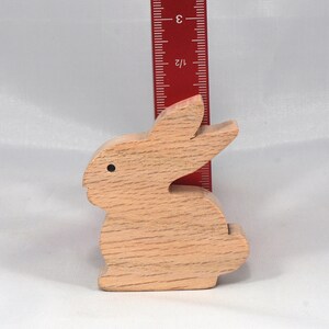 May include: A wooden rabbit figurine, carved from light brown wood, standing approximately 3 inches tall. The rabbit has a simple, rounded design with a small black dot for an eye.
