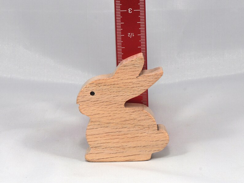 May include: A wooden rabbit figurine, carved from light brown wood, standing approximately 3 inches tall. The rabbit has a simple, rounded design with a small black dot for an eye.