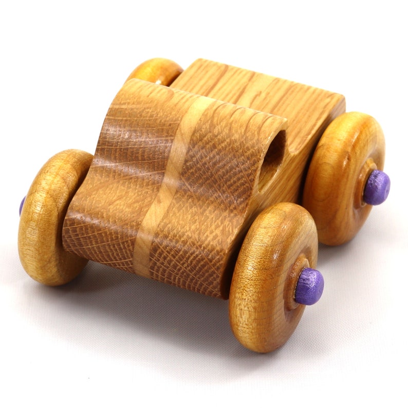 May include: Wooden toy car with a natural wood finish. The car features four large wooden wheels with purple center caps. The body of the car is made of light brown wood with a curved design.