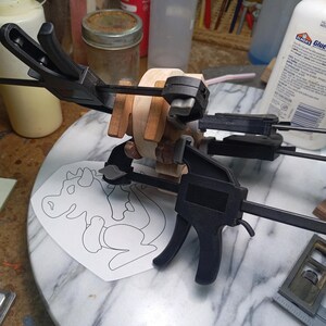 Several clamps hold light and dark wood pieces together as the baby dragon is assembled. A paper drawing of the dragon lies beneath it on the workbench.