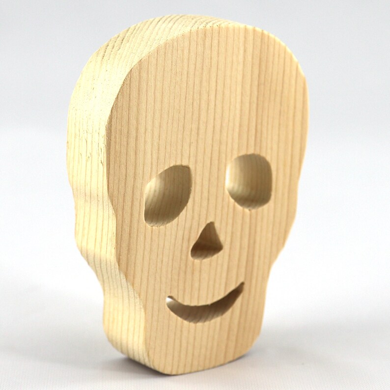 A handcrafted, unfinished light wood Halloween skull cutout featuring carefully carved eye sockets, a nose, and a cheerful, smiling mouth. Displayed upright against a white background.