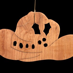 Handmade wooden Christmas tree ornament shaped like a cowboy hat, featuring a sprig of holly tucked into the hat band, star-shaped leaf cutouts, and round berry accents.