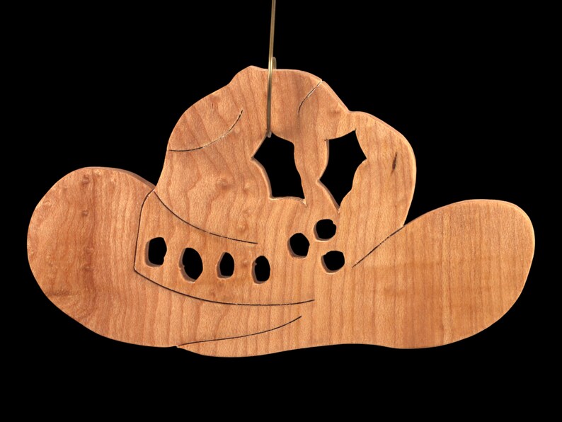 Handmade wooden Christmas tree ornament shaped like a cowboy hat, featuring a sprig of holly tucked into the hat band, star-shaped leaf cutouts, and round berry accents.