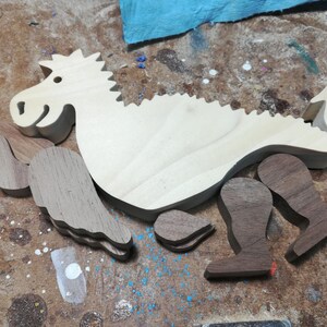 Unassembled baby dragon body with contrasting hardwood wings, legs, arms, and ears arranged on a woodworking bench before final sanding and assembly.