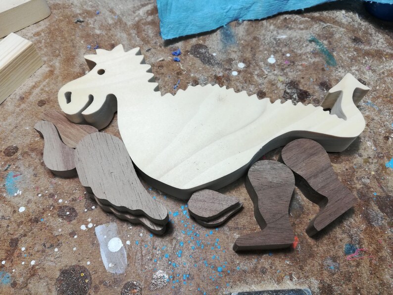 Unassembled baby dragon body with contrasting hardwood wings, legs, arms, and ears arranged on a woodworking bench before final sanding and assembly.