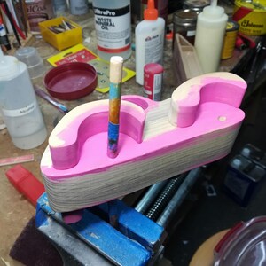 A wooden fat-fender toy car rests on its side above a blue-taped bench vise during painting. Hot pink paint covers much of the exposed side and inner fender curves, while portions of the fenders and the broad body edge remain bare wood. A paint-marked dowel projects vertically from an axle hole and serves as a handle while painting; the wheels are absent. Paint, glue, brushes, and finishing supplies fill the surrounding workbench.