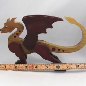 Handmade wooden dragon figurine made from contrasting hardwoods with a light natural wood body and head paired with rich dark reddish-brown wings, legs, and horns. Displayed with a wooden carpenters rule for scale.