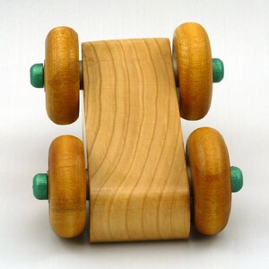 A handmade wooden toy monster truck with a natural wood body and four large wheels. The body and wheels are finished with amber shellac, and the hubs are painted with metallac emerald green acrylic paint. The toy is designed with a simple, classic aesthetic.