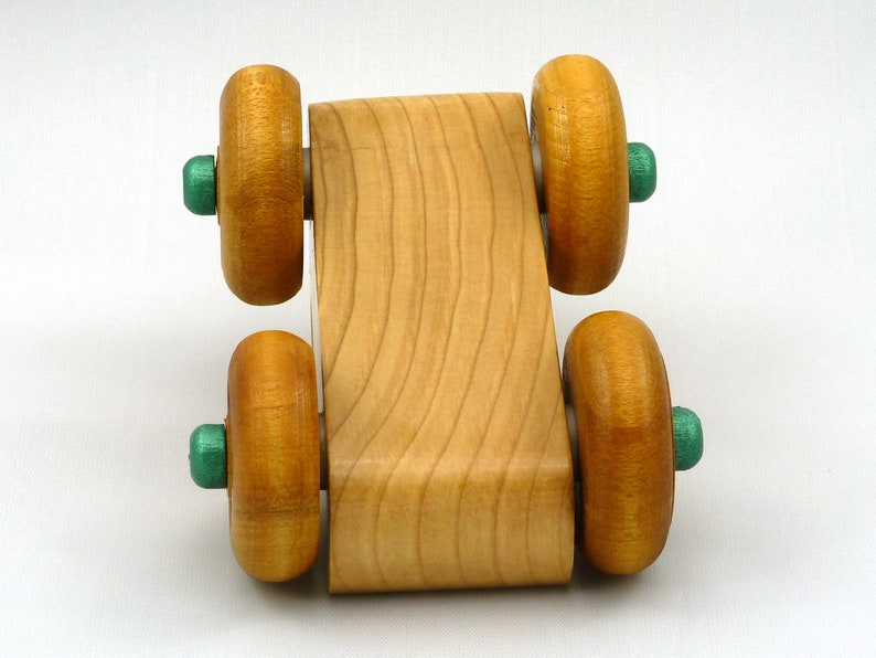 A handmade wooden toy monster truck with a natural wood body and four large wheels. The body and wheels are finished with amber shellac, and the hubs are painted with metallac emerald green acrylic paint. The toy is designed with a simple, classic aesthetic.
