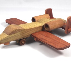 May include: A wooden toy airplane with a tan and reddish-brown color scheme. The toy features wings, a fuselage, and two engine pods. The plane has a propeller and small wheels. The toy is made of wood and appears to be handcrafted.