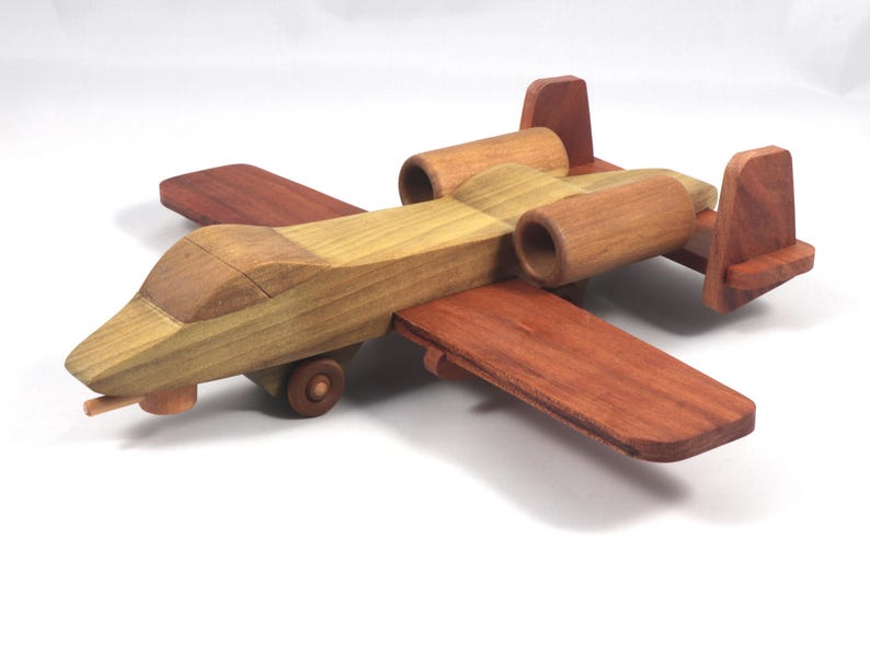 May include: A wooden toy airplane with a tan and reddish-brown color scheme. The toy features wings, a fuselage, and two engine pods. The plane has a propeller and small wheels. The toy is made of wood and appears to be handcrafted.