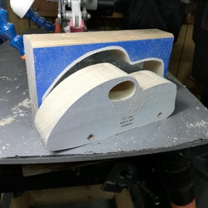 A freshly cut pine toy car body rests on a sawdust-covered scroll saw table, with the matching waste block standing behind it. Blue painter’s tape surrounds the car-shaped opening in the block. The body still carries its paper pattern, with an oval window opening and two drilled axle holes. Exposed wood grain follows the curved roof and sloping rear, showing the fat-fender car before sanding, fender attachment, and painting.