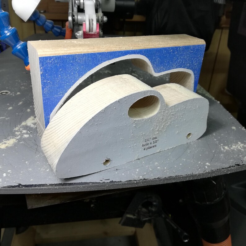 A freshly cut pine toy car body rests on a sawdust-covered scroll saw table, with the matching waste block standing behind it. Blue painter’s tape surrounds the car-shaped opening in the block. The body still carries its paper pattern, with an oval window opening and two drilled axle holes. Exposed wood grain follows the curved roof and sloping rear, showing the fat-fender car before sanding, fender attachment, and painting.
